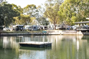 Finding the best caravan parks in Australia, looking over a lake towards the grassy and shaded caravan park spaces on a sunny day