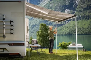 A man is setting up his caravan awning looking out over the lush green grass and lake with mountains in the background