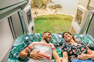 A couple laying on their bed in their RV with the read doors open and a view of the beach behind them